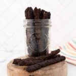 Homemade venison jerky for dogs in a mason jar and 3 pieces on a board in front of it.