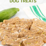 Homemade bone-shaped zucchini dog treats on a plate and a zucchini behind it.