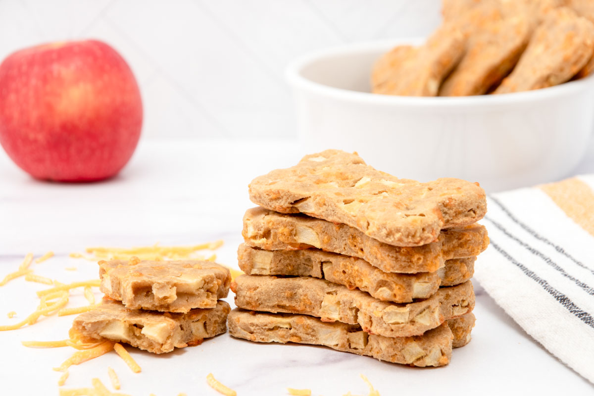 Homemade apple cheddar dog treats in a stack and bowl and an apple.