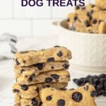 Homemade bone-shaped blueberry dog treats in a stack and in a bowl behind it.