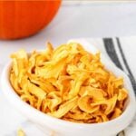 Homemade dehydrated pumpkin dog treats in a small oval dish and a pumpkin behind it.