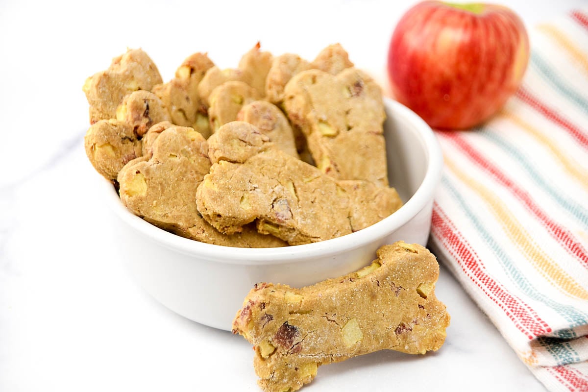 Homemade dog treats with apple and bacon in a bowl and an apple beside it.